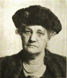 B&W portrait photo of a woman wearing a dark hat, pearls, and dark jacket