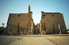 Par trapezoidnih kamnitih pilonov spremlja prehod, skozi katerega je vidna vrsta stebrov. Pred pilonoma je nekaj velikih kipov in obelisk.