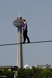 A man with a purple shirt and black pants tightrope walks with a large tower in the background