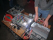 A photo of Pete Swanson (only lower part of his body is visible) performing live, with various pieces of electronic equipment placed on a table in front of him.