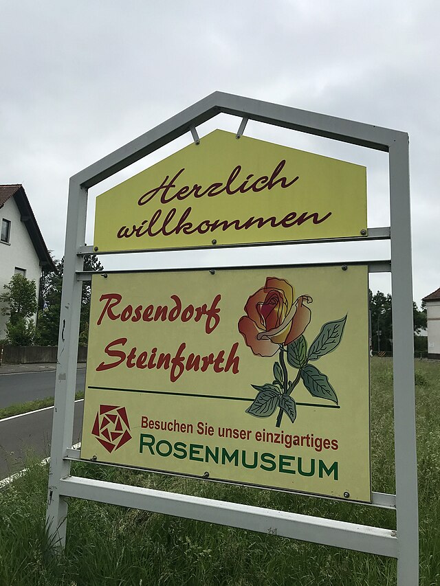 Ortseingangsschild vom Rosendorf Steinfurth heißt seine Gäste willkommen und ruft zum Besuch des einzigartigen Rosenmuseums auf.