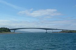 A body of blue water is spanned by a concave bridge of modern design in the middle distance. A small lighthouse can be seen beyond the bridge under its span.