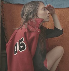 A white woman is sitting perpendicular to the camera, facing to the right; she has a red jacket (emblazoned with "05") draped over her shoulders, and her left hand is shielding her eyes.