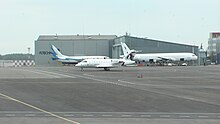 Seitliche Farbfotografie von drei weißen Flugzeugen auf dem Flugfeld. Das rechte Flugzeug ist größer. Im Hintergrund sind graue Gebäude.