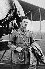 Half portrait of young dark-haired man in military uniform with coat over left arm, standing in front of biplane