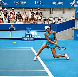 Tennispelaaja vaaleansinisessä mekossa taivuttaa polvea lyömään matalaa palloa kädellä molemmin käsin.