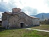 A medieval church constructed of stone and brickwork with the entrance and dome visible standing in a grassy area with wooded mountains in the background