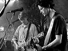 Two men holding guitars onstage. Man at left is looking downwards, right hand strummings strings, left hand on fret board. Second man is half turned with his left hand high on the fret board.