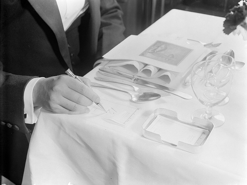 File:Gedekte tafel met bestek en een menu de hand van een man die '12' heeft geschre, Bestanddeelnr 190-0709.jpg