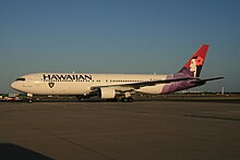 A white twin-engine plane painted with the word "HAWAIIAN" in the front above the windows, a black, silver, and white logo of a pirate's head on the forward bottom of the fuselage, and a woman in different purple hues on the tail taxis at an airport