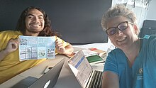 two people smiling faces with a desk and computer and a little unflded zine. kasi has wavy brown hair, Lisa has glasses and short gray hair