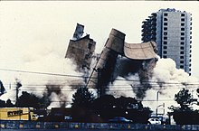 The Alfred P. Murrah building is being demolished, and the image shows the building in mid-collapse. A Ryder truck is visible at the bottom left, and the Regency Towers building can be seen in the background at the far right. The demolition has created large clouds of dust that take up a portion of the image.