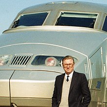 Cropped colour photograph of man in dark jacket, shirt and tie standing in front of French TGV high-speed train.