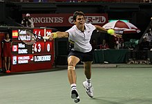 Raonic lunging to his right behind the baseline, with his racquet stretching to reach the approaching ball. Scoreboard in the background.