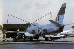 A monoplane aircraft with three men and a fuel tanker truck. One of the men is wearing military uniform and the other two are clad only in shorts. The aircraft is mainly painted grey, but is marked with the Royal Australian Air Force roundel and stripes on its tail. The tail of another aircraft of similar appearance is visible in the background.
