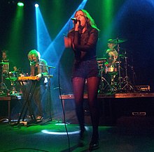 A woman wearing a black top and blue shorts performs in a dark room with her band behind