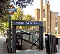 A green sign with the words "WARWICK AVENUE STATION" written in white letters on a dark blue rectangle all under a light blue sky with white clouds