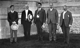Photographie en noir et blanc de cinq hommes en costume posant chapeau à la main