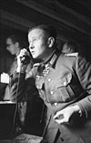 Black-and-white portrait of a man on the phone in semi profile wearing a military uniform with an Iron Cross displayed at his neck. He is holding a cigarette in his left hand.
