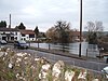 In the foreground are a stone wall and road. Beyond is an area of water surrounded by trees and white fronted houses.