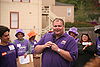 A man in a purple shirt uses a microphone to speak to a crowd outside.