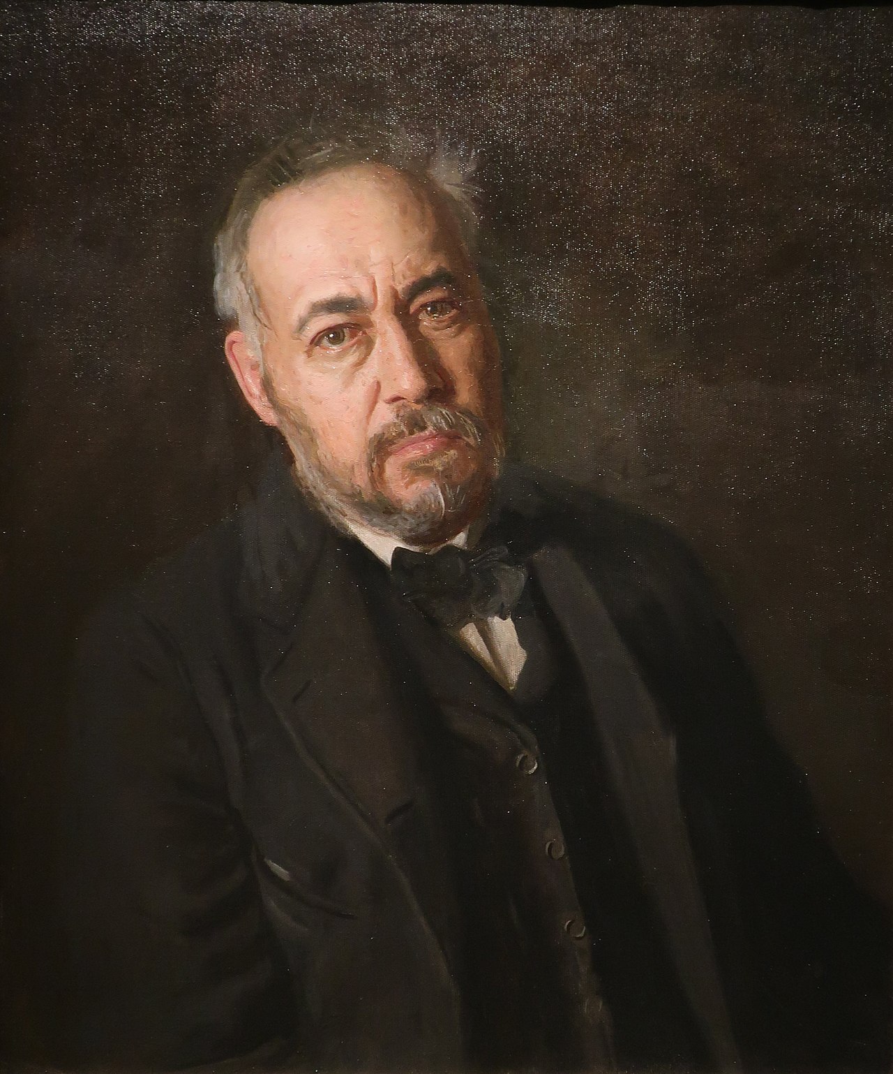 A man, in front of a solid dark background and wearing a dark suit with a dark bow tie, with greying hair, a trim beard and moustache, stares out from dark brows directly at the viewer.