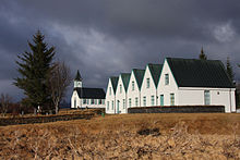 Petit bâtiment blanc à toit vert avec cinq pignons, et une église aux couleurs similaires plus en arrière.