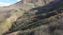 View of grass-covered hillside and trail at the crash site of the 2020 Calabasas helicopter crash. In the center of the photograph, debris from the helicopter crash can be seen, along with investigators working amongst the wreckage.