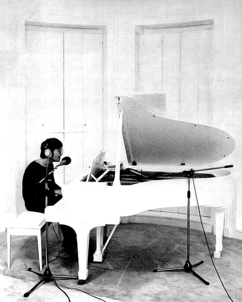 A black and white photo of Lennon sitting at a white parlour grand piano. He is wearing headphones and a dark shirt.