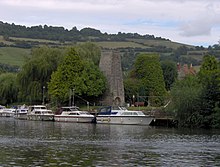 Looking across water to moored boats. Beyond them is a stone chimney surrounded by trees, with hills in the distance.