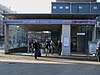 Sotto un cartello blu con la scritta "STAZIONE DELLA METROPOLITANA DI KING'S CROSS ST. PANCRAS diverse persone escono da un ingresso che conduce a una scalinata. A destra ci sono delle porte d'argento sotto un cartello con la scritta "Ascensore". Il cielo è coperto, sopra l'ingresso è buio edificio temporaneo blu