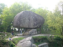 Rocha de grande porte equilibrada em um pequeno pedestal.  Alguns visitantes dão uma ideia da escala: vários metros de altura.