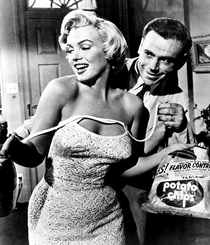 Monroe in The Seven Year Itch. She is holding a bag of chips and wearing a dress, which shoulder straps are undone. Behind her is Tom Ewell, who is holding the straps.