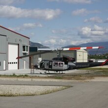 A blue and red helicopter sits just off an airport runway, with a building shown in the background.