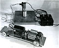The important elements of this system are the clutches housed in the twin steel cylinders on the chassis of the model, the control box at the top right, and the pivitol connector between the control box and the motor and clutches of the model (top center). The motor shaft on the model drives a friction disc connected to a small magnetic fluid cltch geared to the rear wheels. One end of the second clutch, housed in the cylinder nearest the rear right wheel, is locked to the frame so that it can be used as a brake. Jacob Rabinow invented the Magnetic Particle Clutch in 1947 during his work on ordnance at the National Bureau of Standards. The particle clutch used a mixture of light lubricating oil and powdered carbonyl iron inside a chamber connecting two free spinning plates. When an electromagnet attached to the chamber was energized, the iron particles would magnetize and attract each other, producing an almost solid mass, which locked the plates together. Controlling the magnetic flux would control the amount of torque between the plates. Because Rabinow invented the Magnetic Particle Clutch as part of his work at the National Bureau of Standards, the United States Government owned the U.S. patent. However, in partnership with his brother Joseph, Rabinow did file for patents in 22 foreign countries. Due of the simple design, precise torque control, smooth operation, and long life, the device found wide application. It was used in Renault and Subaru automobiles in Europe and Japan, in airplane controls, and in the disc file of the IBM RAMAC computer. Reference: Inventing for Fun and Profit. Jacob Rabinow. San Francisco Press, San Francisco, CA. (1990). pp. 49-55.