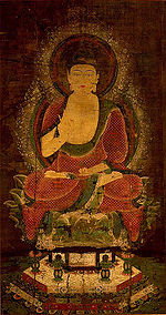 Vista frontale di una divinità seduta a gambe incrociate su un piedistallo.