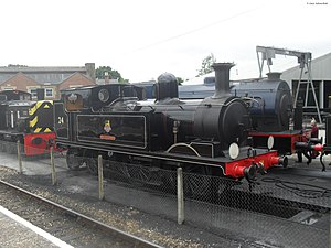 A steam locomotive in a maintenance depot. Beneath it is an inspection pit. To the left is a Diesel shunting locomotive; to the right is another steam locomotive and a crane.