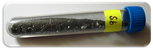 Un vial transparente que contiene pequeños trozos de un sólido negro ligeramente brillante, con la etiqueta "Sb".