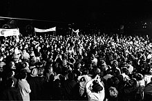 Photo en noir et blanc d'une foule photographiant et applaudissant des personnes en tenue militaire, qui sont au centre.