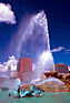 A fountain spouting water from several points with several tall buildings and a blue sky in the background.