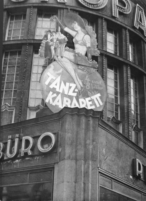A black and white picture of the exterior of a Weimar-era cabaret. The exterior features a cardboard or paper mache figure of a cabaret dancer in a black bra and short skirt.