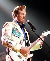 A photograph of a caucasian man with tall red hair and red beard, wearing a white, dazzling jacket and blue undershirt, playing a guitar.