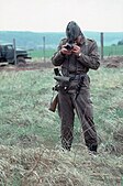 Armed East German border guard standing in a grassy field taking a photograph of the photographer. A border fence and a truck are visible in the background, some distance behind the soldier.