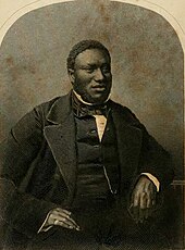 Photographie noir et blanc, trois-quart face d'un homme noir corpulent, assis, un bras posé sur une table, habit et gilet sombre sur chemise blanche