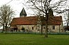 Een stenen kerk met rode pannendaken gezien vanuit het zuiden, met het koor, de zuidelijke zijbeuk en veranda, en de toren met een piramidevormig dak