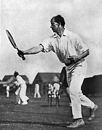 A man in white pants and shirt swings a wooden racket on a grass court, with other male and female tennis players in the background