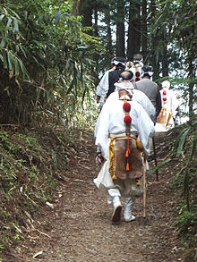 Monks with cropped hair and walking staves hiking up a steep mountain trail in white robes.