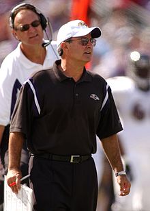 Candid photograph of Zauner standing in front of Brian Billick on a football sideline wearing a black polo shirt and white baseball cap both of which bear the Baltimore Ravens logo