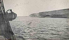 A barren and featureless headland, observed from the side of a ship across a stretch of calm sea