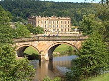 Photo d'un château entouré de bois, avec, au premier plan le pont de pierre à deux arches permettant d'y accéder.